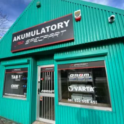 Fasada budynku z blachy trapezowej w kolorze morskim, z szyldem 'Akumulatory Specpart' nad wejściem i oknami wystawowymi prezentującymi akumulatory marek TAB, Exide, Gram i Varta. W oknach naklejki...