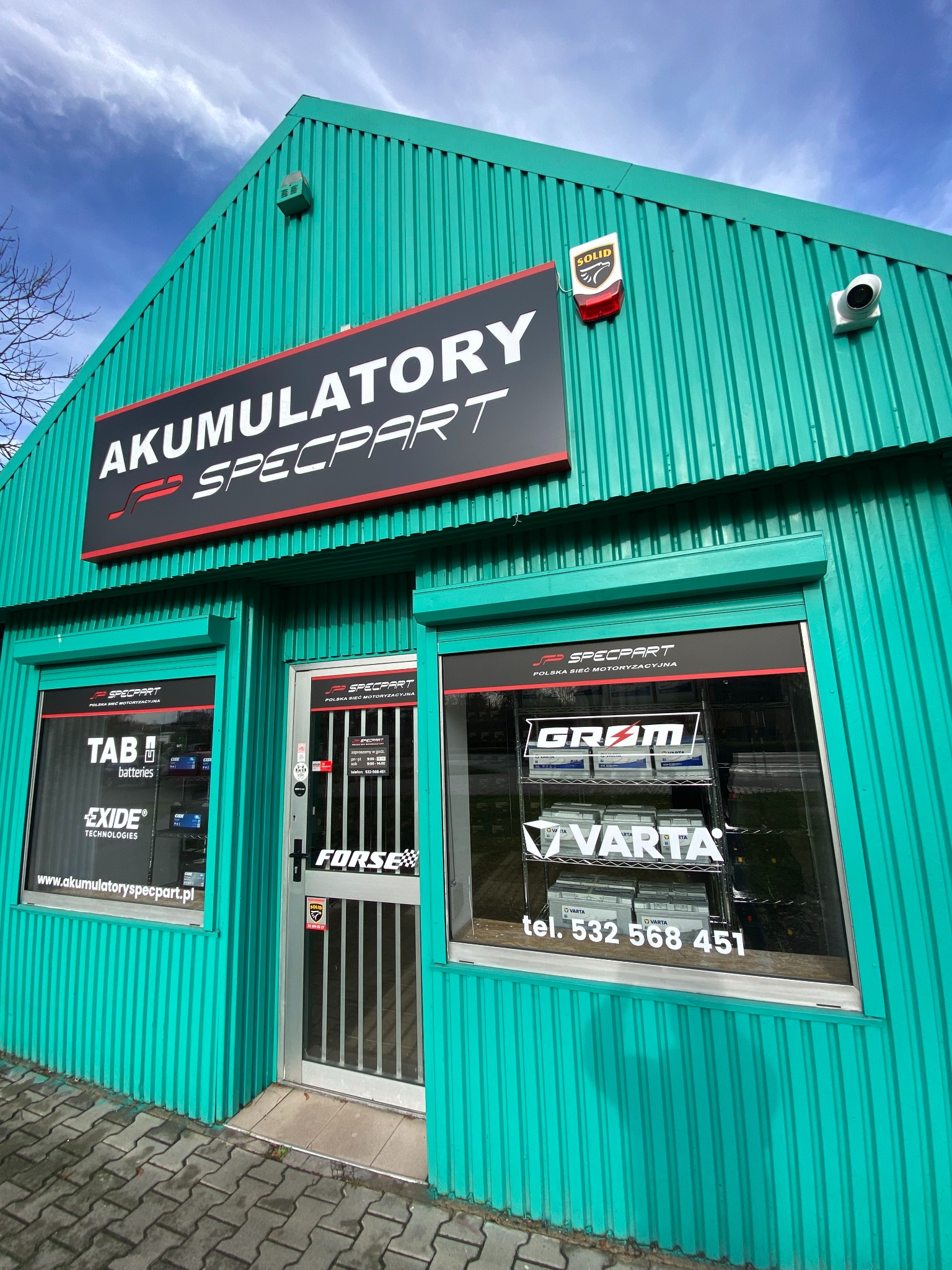 Fasada budynku z blachy trapezowej w kolorze morskim, z szyldem 'Akumulatory Specpart' nad wejściem i oknami wystawowymi prezentującymi akumulatory marek TAB, Exide, Gram i Varta. W oknach naklejki...