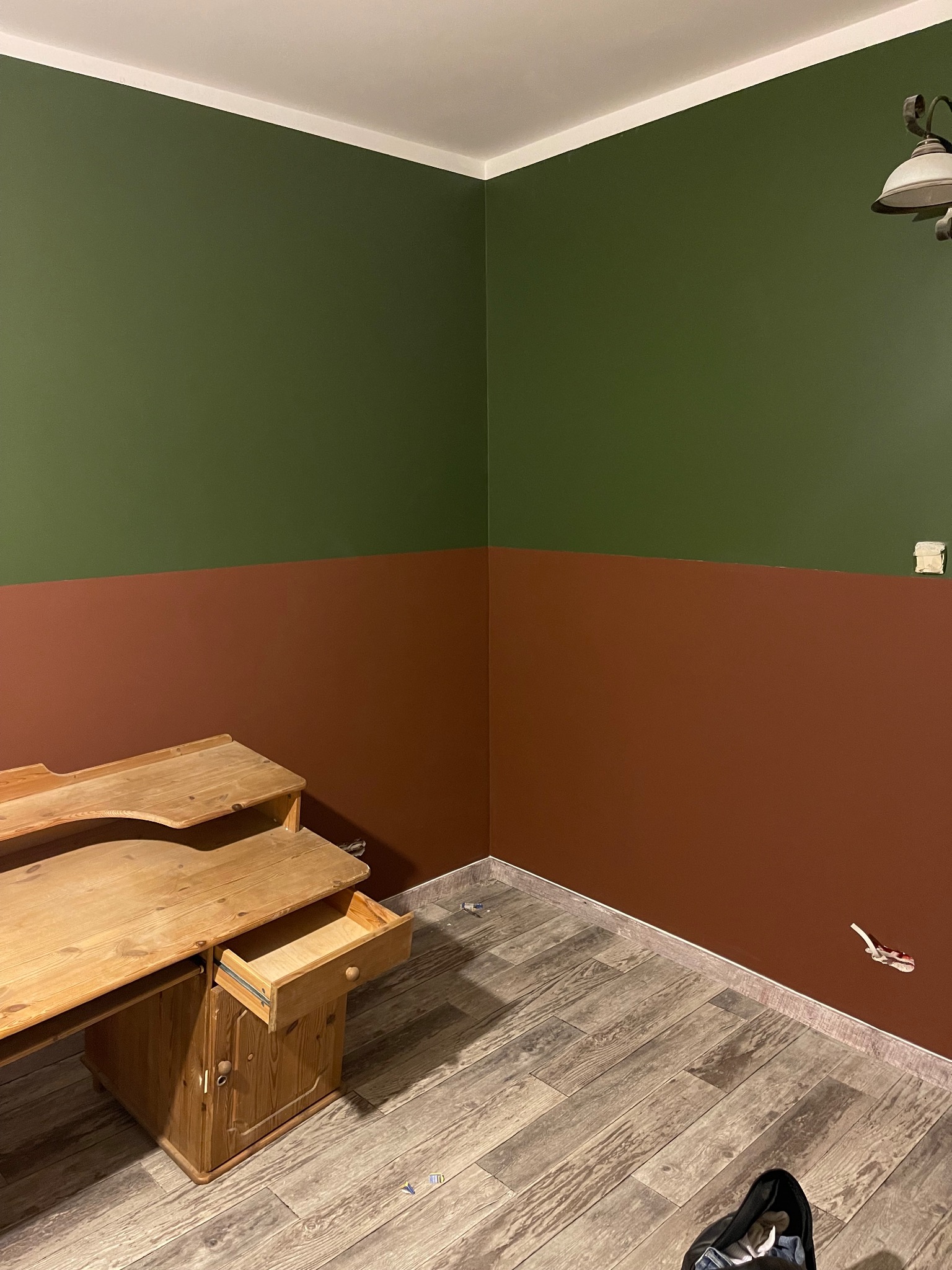 Pokój po malowaniu dwukolorowym: górna część ściany oliwkowozielona, dolna część brązowa, drewniane biurko z wysuniętą szufladą, podłoga z paneli imitujących drewno, widoczne ślady po remoncie.