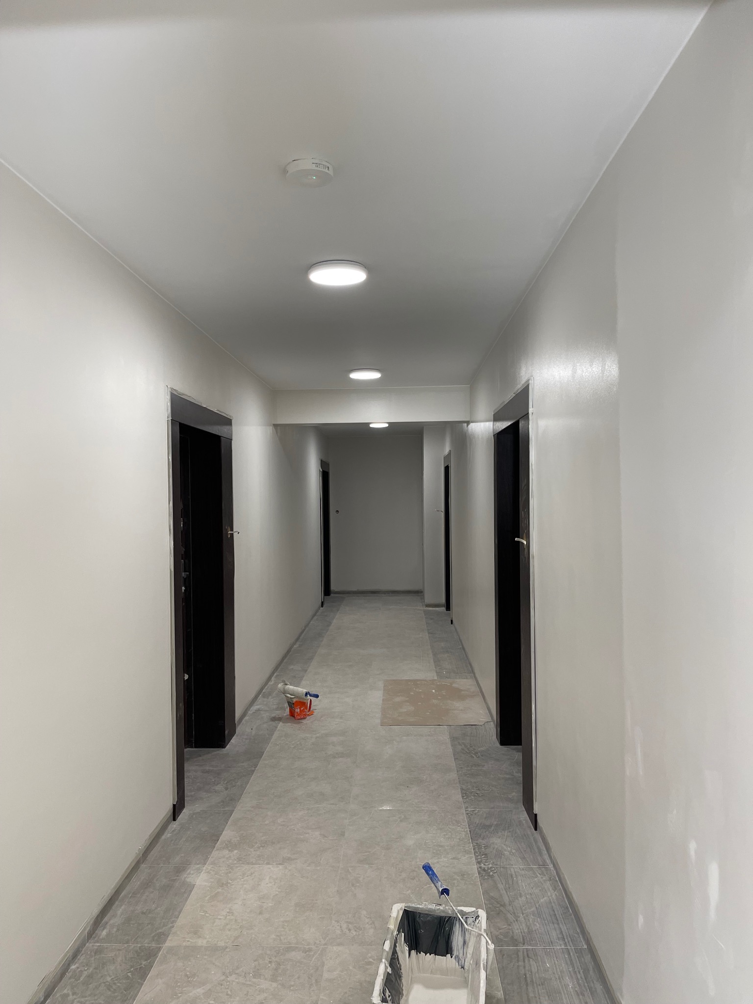 Korytarz w trakcie malowania na biało, widoczne narzędzia malarskie: kuweta z farbą, wałek, pędzel oraz ślady świeżo pomalowanej ściany, ciemne futryny drzwi kontrastują z jasnymi ścianami.