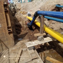 Hydro gadżet - Wykop z widocznymi rurami wodociągowymi w kolorze niebieskim i gazowymi w kolorze żółtym, zabezpieczony stalową szalunką i drewnianymi podporami. Widoczny fragment ulicy w tle.