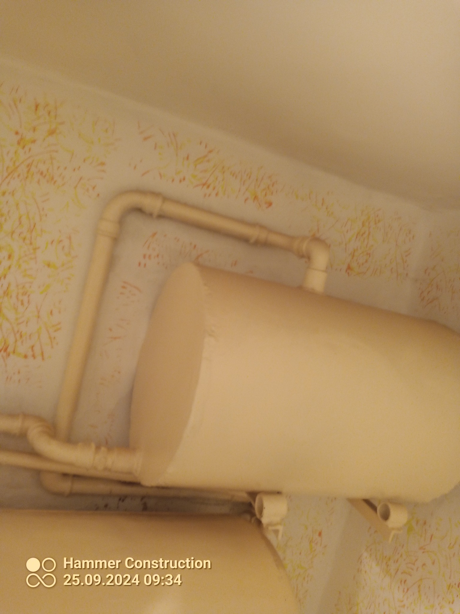 Kremowy, poziomy zbiornik z rurami przymocowany do ściany z wzorzystą tapetą, widoczne mocowania zbiornika.