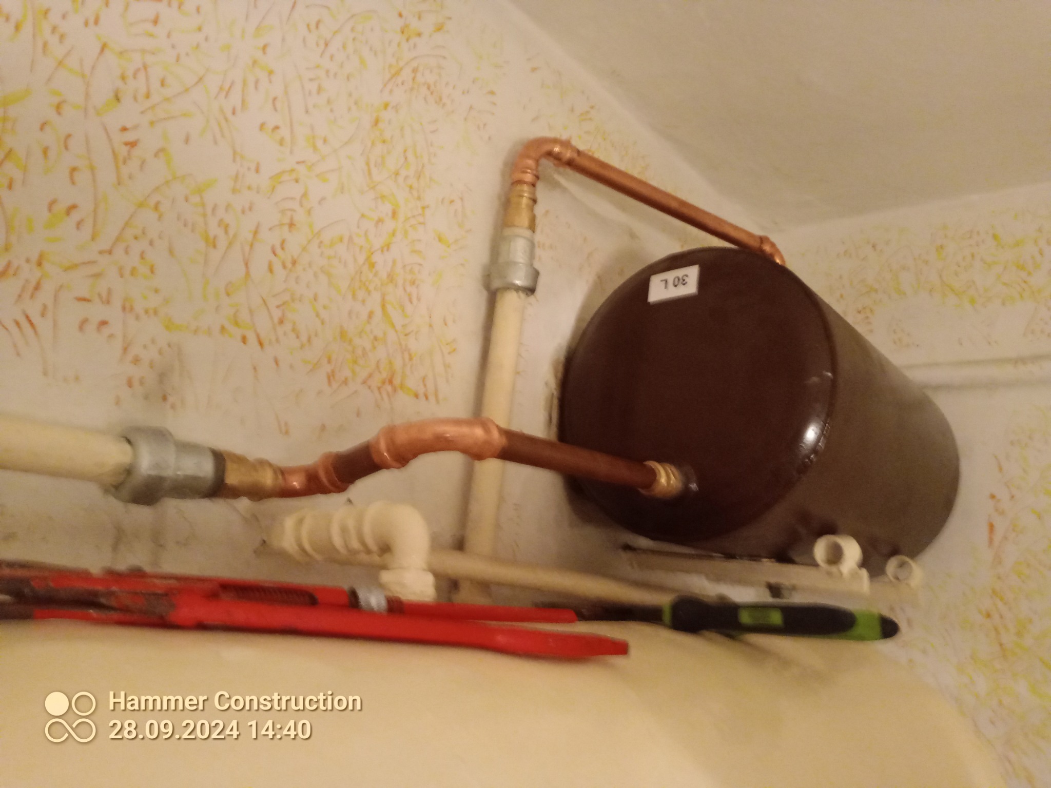 Brązowy bojler 30L podłączony miedzianymi rurami do instalacji, widoczny klucz nastawny i śrubokręt na tle ściany z wzorzystą tapetą.
