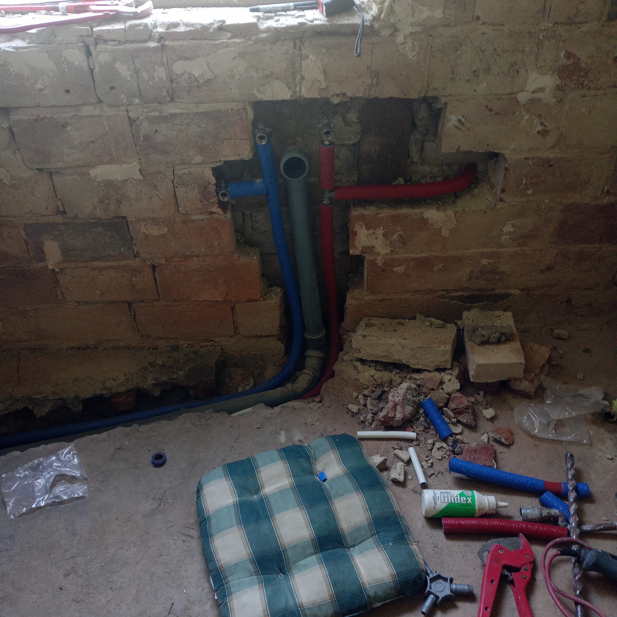 Fragment ściany z cegły z nową instalacją hydrauliczną: rury w kolorze niebieskim, szarym i czerwonym, narzędzia hydrauliczne i poduszka w kratę na podłodze.