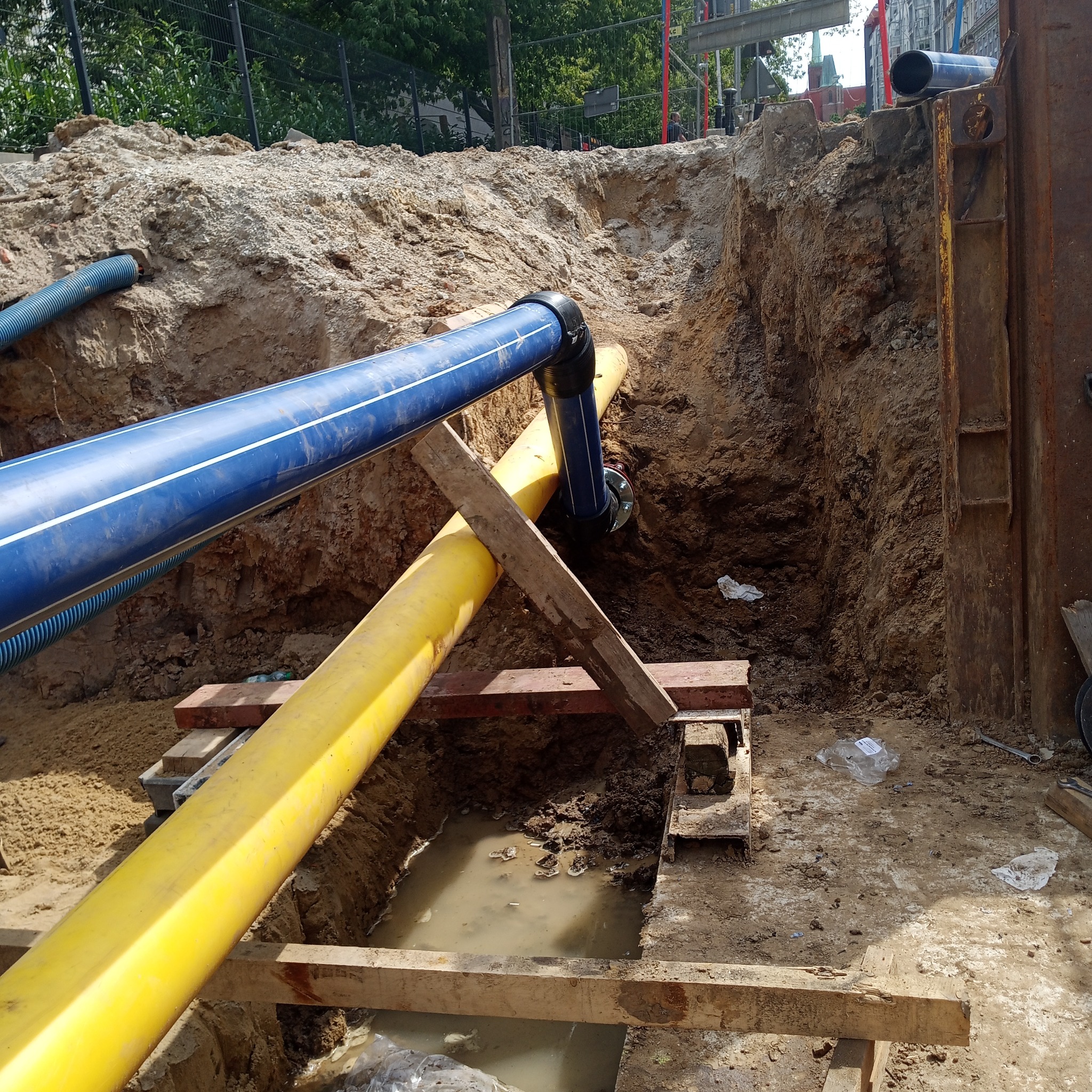 Wykop z widocznymi rurami: niebieską z białymi paskami i żółtą, podpartymi drewnianymi belkami, w tle ziemia i stalowe zabezpieczenia wykopu.