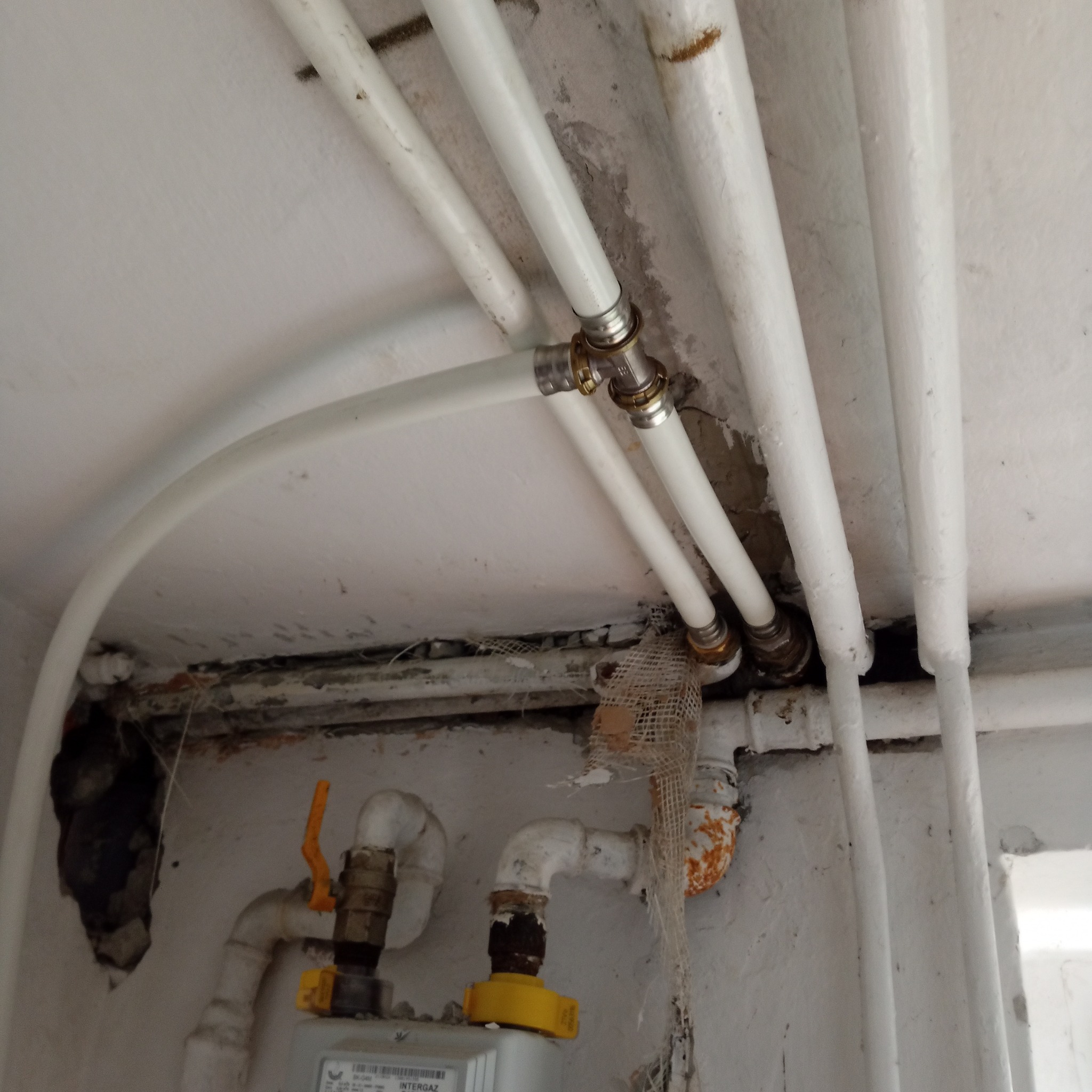 Białe rury wodne z mosiężnymi złączkami biegnące wzdłuż ściany i sufitu, widoczne ślady po naprawach i niedoskonałości na ścianie i suficie, stary gazomierz w tle.