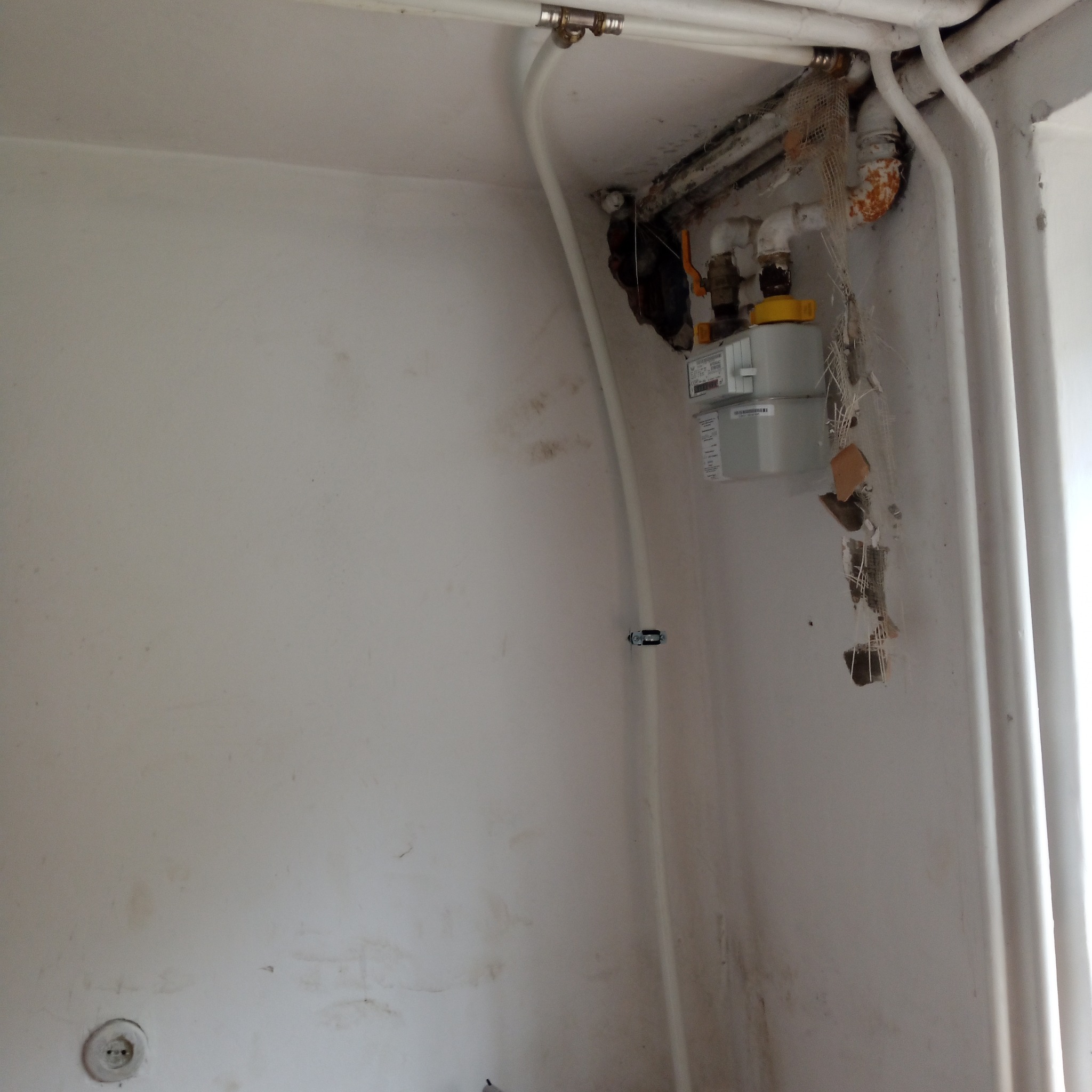 Białe rury instalacji wodnej i gazomierz w rogu pomieszczenia z widocznymi śladami po kuciu w ścianie.