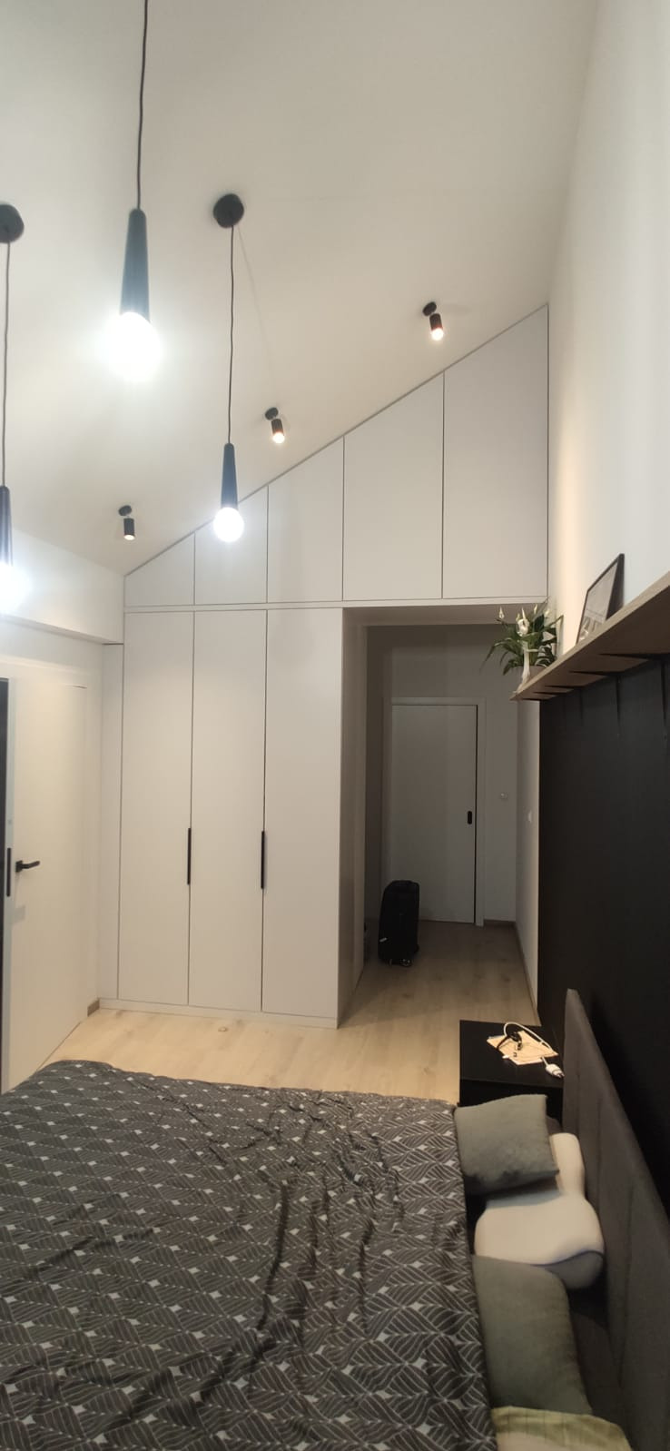 Biała zabudowa szafy wnękowej z czarnymi uchwytami w sypialni ze skośnym sufitem i wiszącymi lampami.