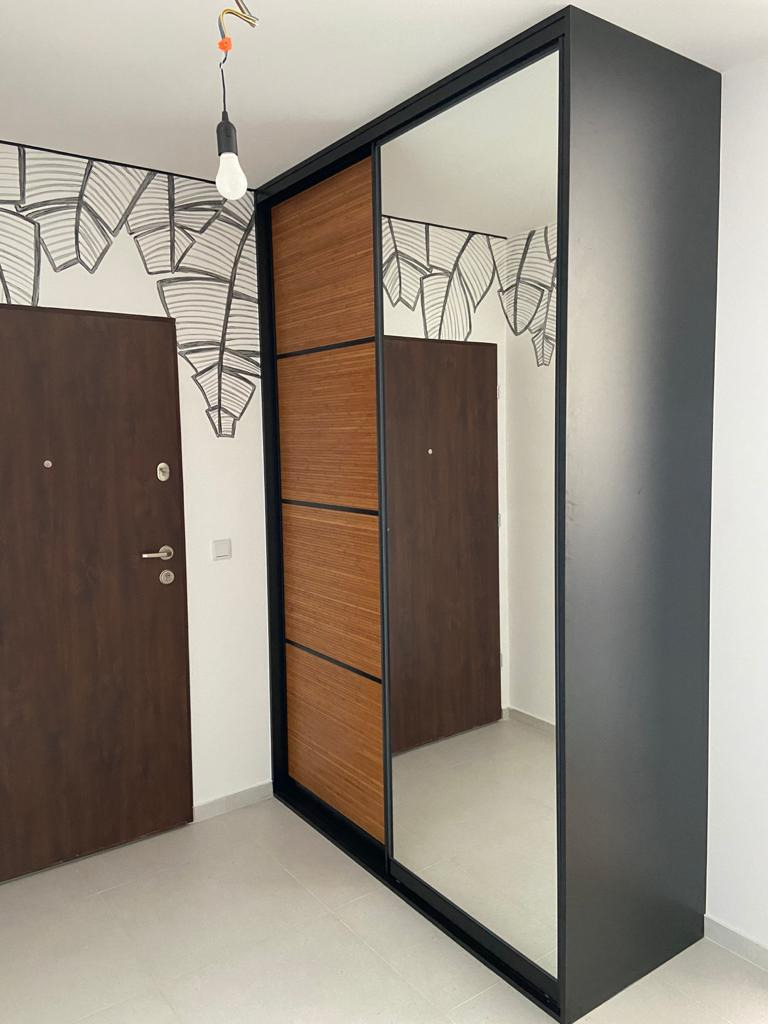 Nowoczesna szafa z przesuwnymi drzwiami: czarna rama, drewniane panele i lustro, widoczna na tle ściany z graficznym motywem liści i brązowymi drzwiami.