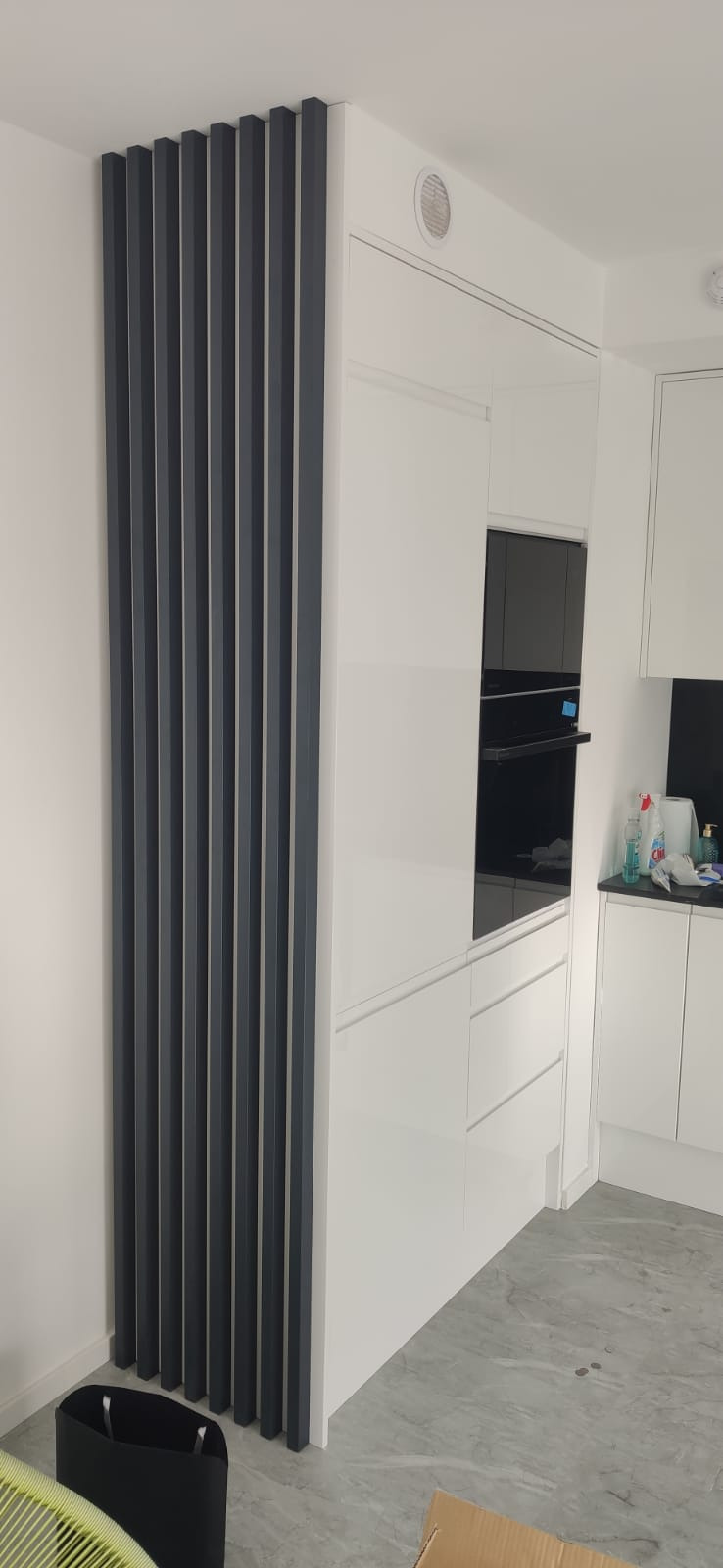Nowoczesna kuchnia na wymiar w Siechnicach z białymi frontami i czarnym piekarnikiem w zabudowie, uzupełniona o dekoracyjny panel z pionowych, szarych listew.