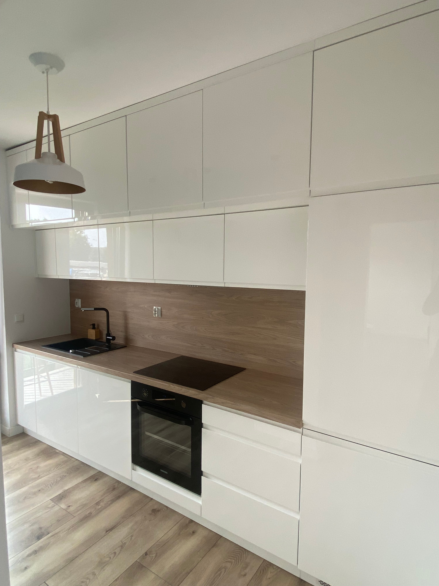 Nowoczesna, biała kuchnia z drewnianym blatem i panelem ściennym, oświetlona wiszącą lampą z elementami drewna, widok na płytę indukcyjną i piekarnik.