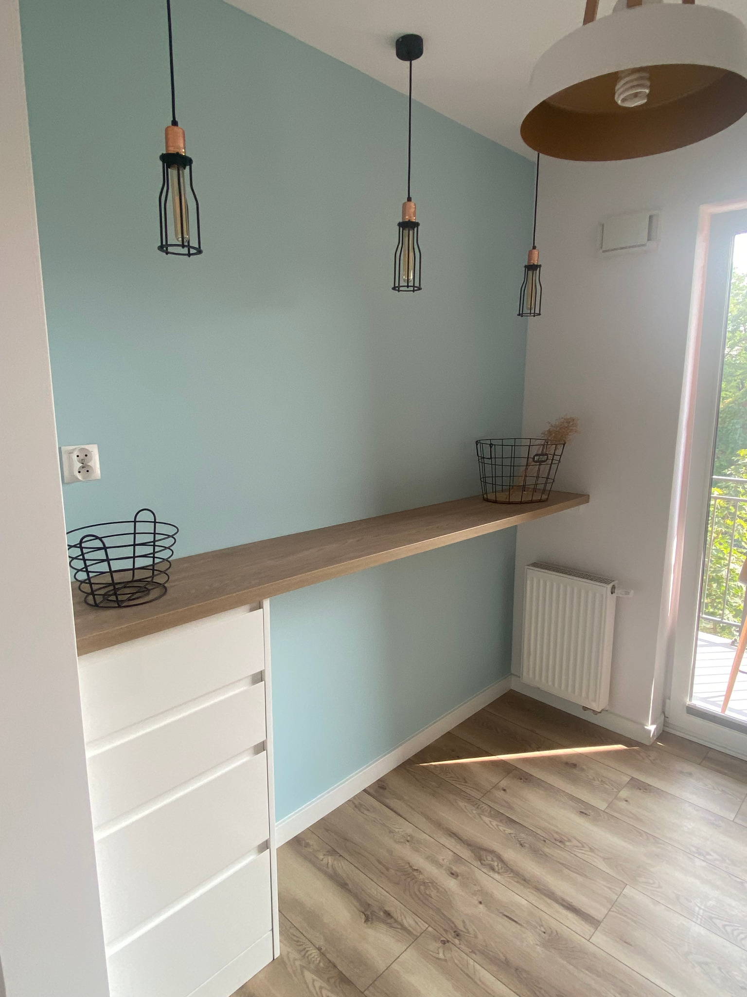 Nowoczesne wnętrze z blatem z jasnego drewna zamontowanym na ścianie w kolorze pastelowej mięty, pod nim biała komoda z szufladami, nad blatem wiszące lampy z czarnymi kloszami, na blacie druciane...