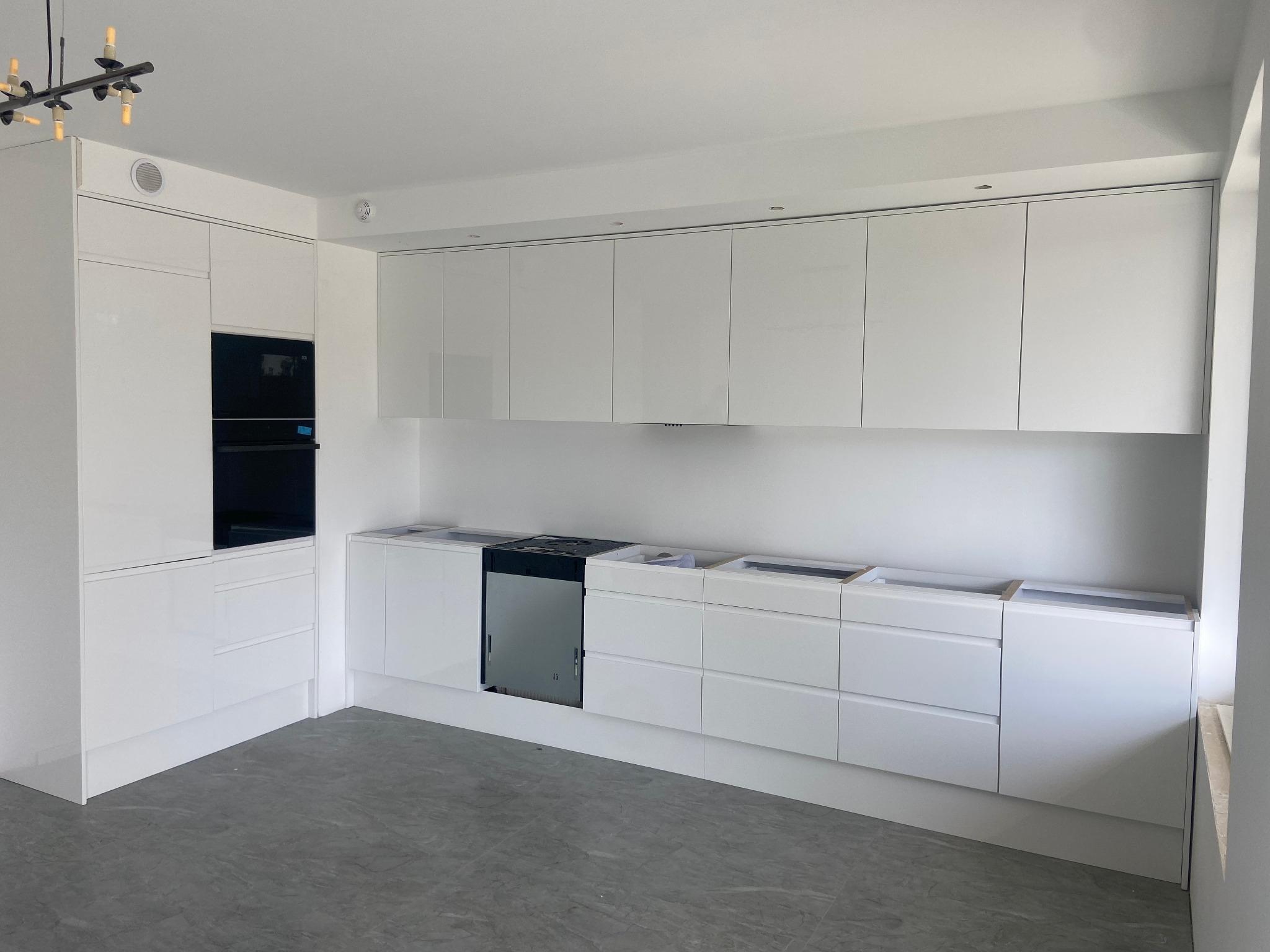 Nowoczesna, biała zabudowa kuchenna z błyszczącymi frontami, jeszcze bez blatów, widoczny piekarnik w zabudowie i płyta indukcyjna w trakcie montażu, szara podłoga.