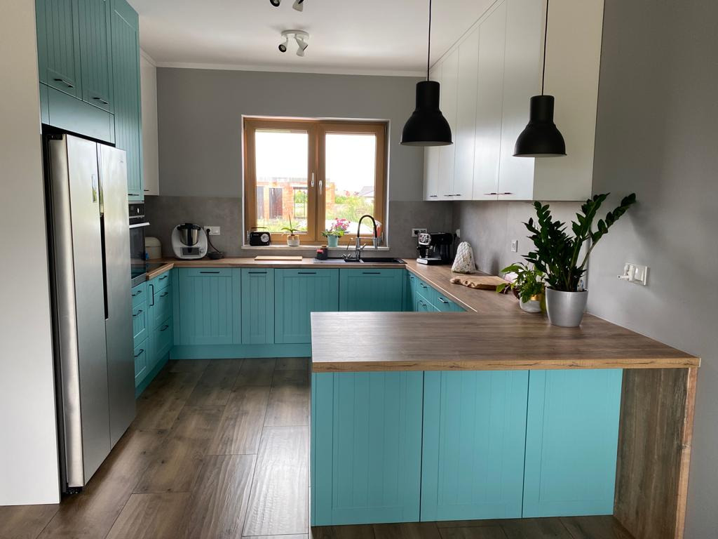 Jasna kuchnia w stylu nowoczesnym z turkusowymi frontami szafek, drewnianym blatem i wyspą kuchenną, oświetlona czarnymi lampami wiszącymi.