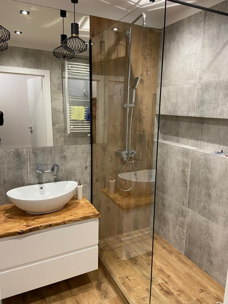 Nowoczesna łazienka z drewnianymi akcentami: blat umywalki z surowego drewna, kabina prysznicowa typu walk-in z drewnianą ścianą, szara płytka na ścianach i podłodze, wiszące lampy z czarnymi...