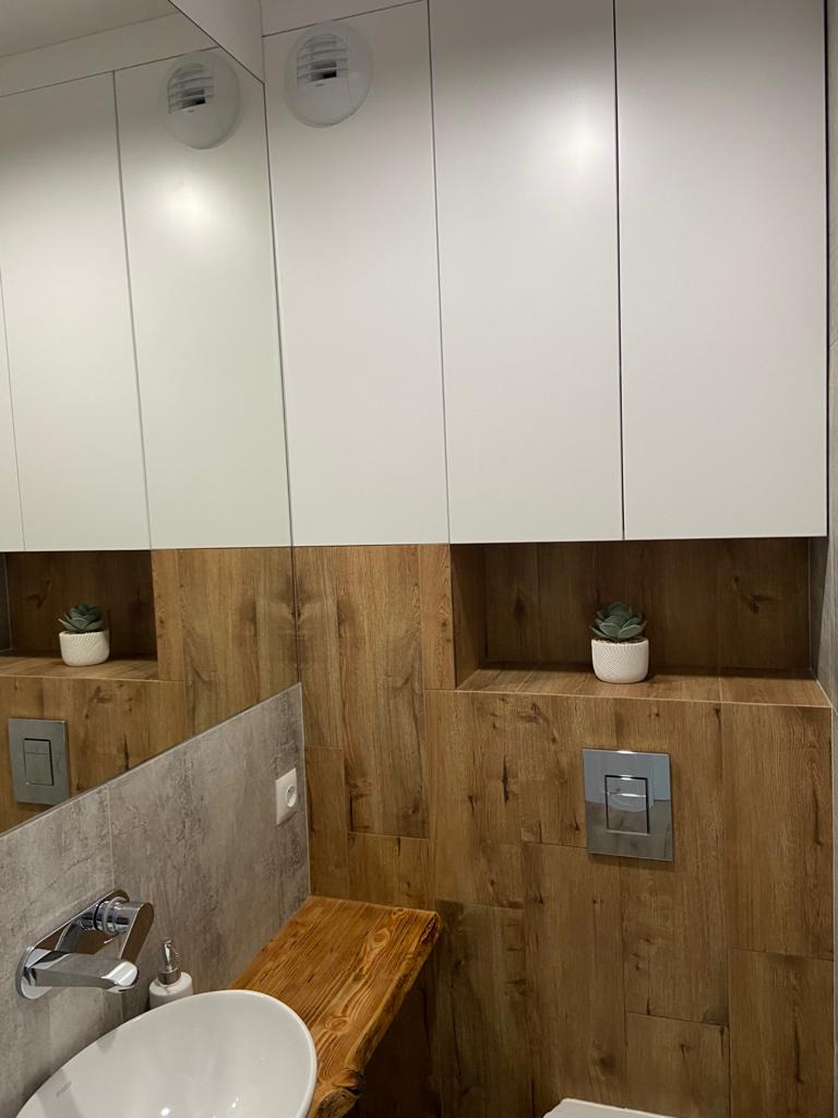Nowoczesna łazienka z drewnopodobnymi płytkami, białymi szafkami wiszącymi z lustrem, drewnianym blatem pod umywalką i rośliną doniczkową w niszy.