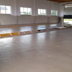 Świeżo wylana, mokra posadzka w hali przemysłowej, z widocznymi drewnianymi szalunkami i laserowym niwelatorem ustawionym na środku, okna w tle.