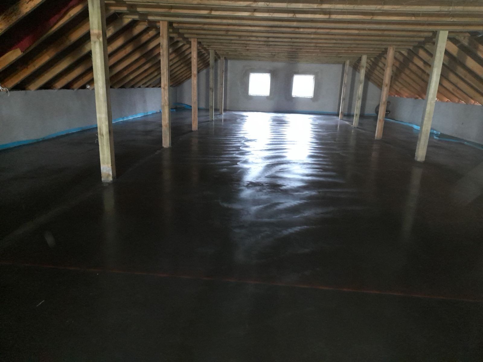 Świeżo wylana, ciemna posadzka przemysłowa w surowym stanie, z drewnianymi słupami podtrzymującymi dach na poddaszu, widoczne dwa okna w oddali.