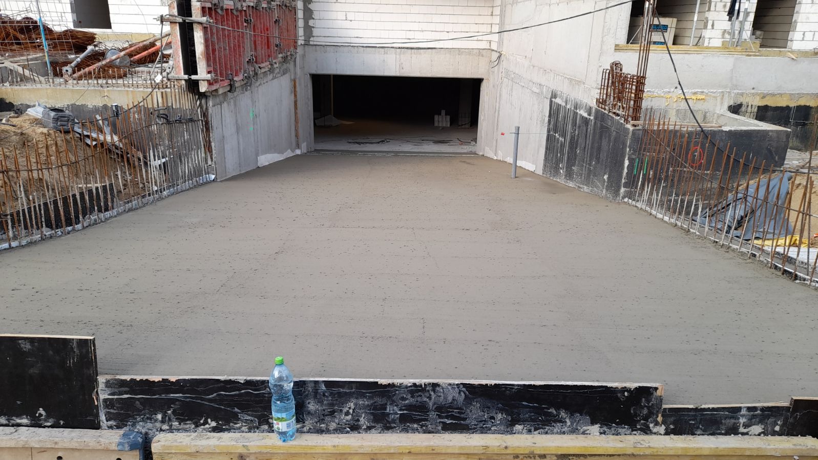 Świeżo wylany beton na przygotowanym podłożu z widocznym zbrojeniem i szalunkami, butelka wody mineralnej w dolnym lewym rogu.