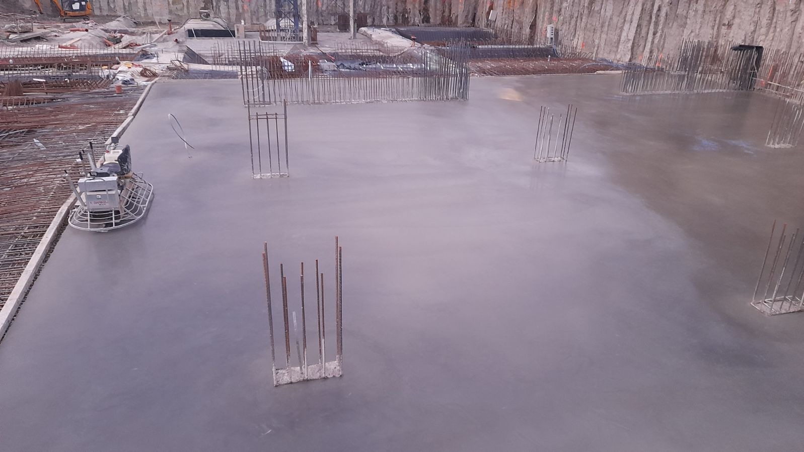 Świeżo wylana, gładka betonowa posadzka w trakcie obróbki zbrojona wystającymi prętami; widoczna maszyna do zacierania betonu oraz zbrojenia w tle.