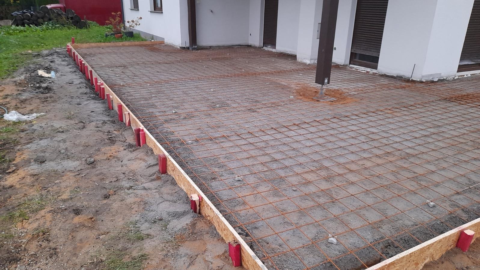 Przygotowanie podłoża pod wylewkę: szalunek z płyty OSB, czerwone kołki stabilizujące i siatka zbrojeniowa na utwardzonym gruncie przed budynkiem.