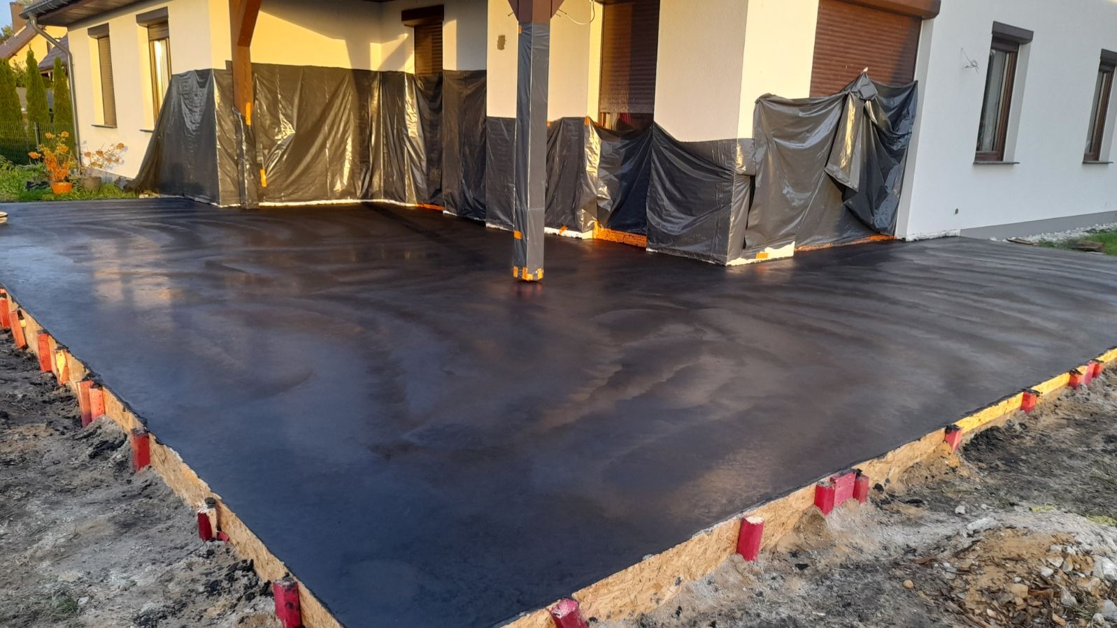 Wylana, świeża, ciemnoszara posadzka na zewnątrz budynku jednorodzinnego, zabezpieczonego czarną folią budowlaną, z widocznym szalunkiem z czerwonymi kołkami.