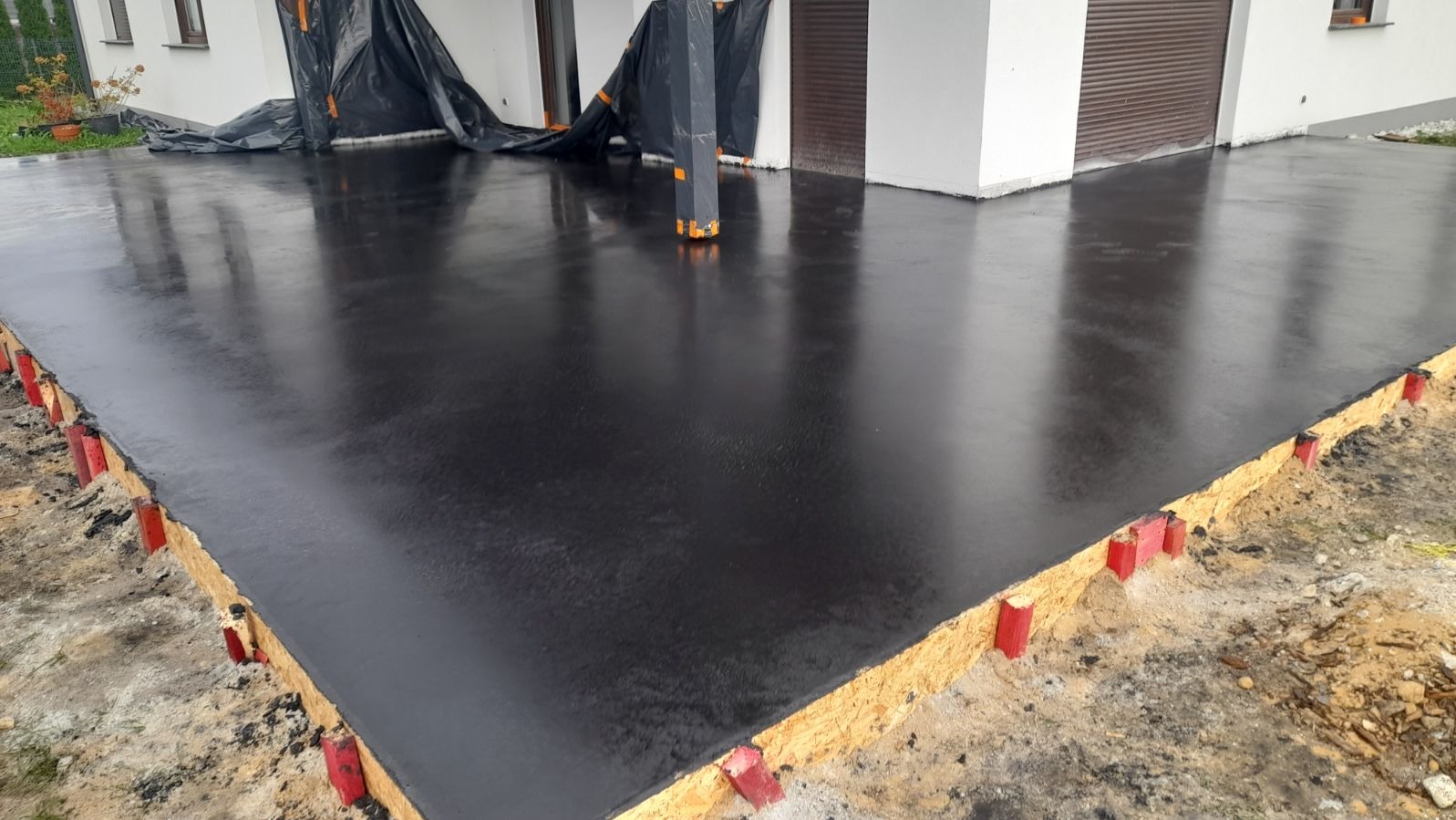 Świeżo wylana, błyszcząca, czarna posadzka z deskowaniem i czerwonymi kołkami stabilizującymi, częściowo zakryta folią ochronną, w tle budynek z roletą.