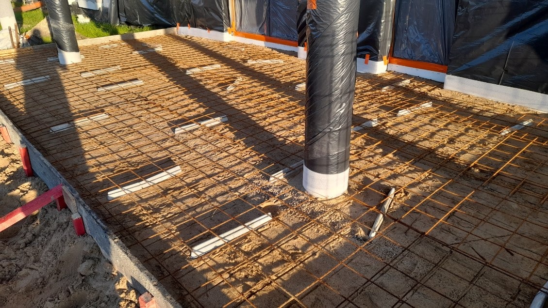 Przygotowanie podłoża pod wylewkę: siatka zbrojeniowa na podkładkach dystansowych, otuliny słupów zabezpieczone folią, widoczne szalunki obwodowe.