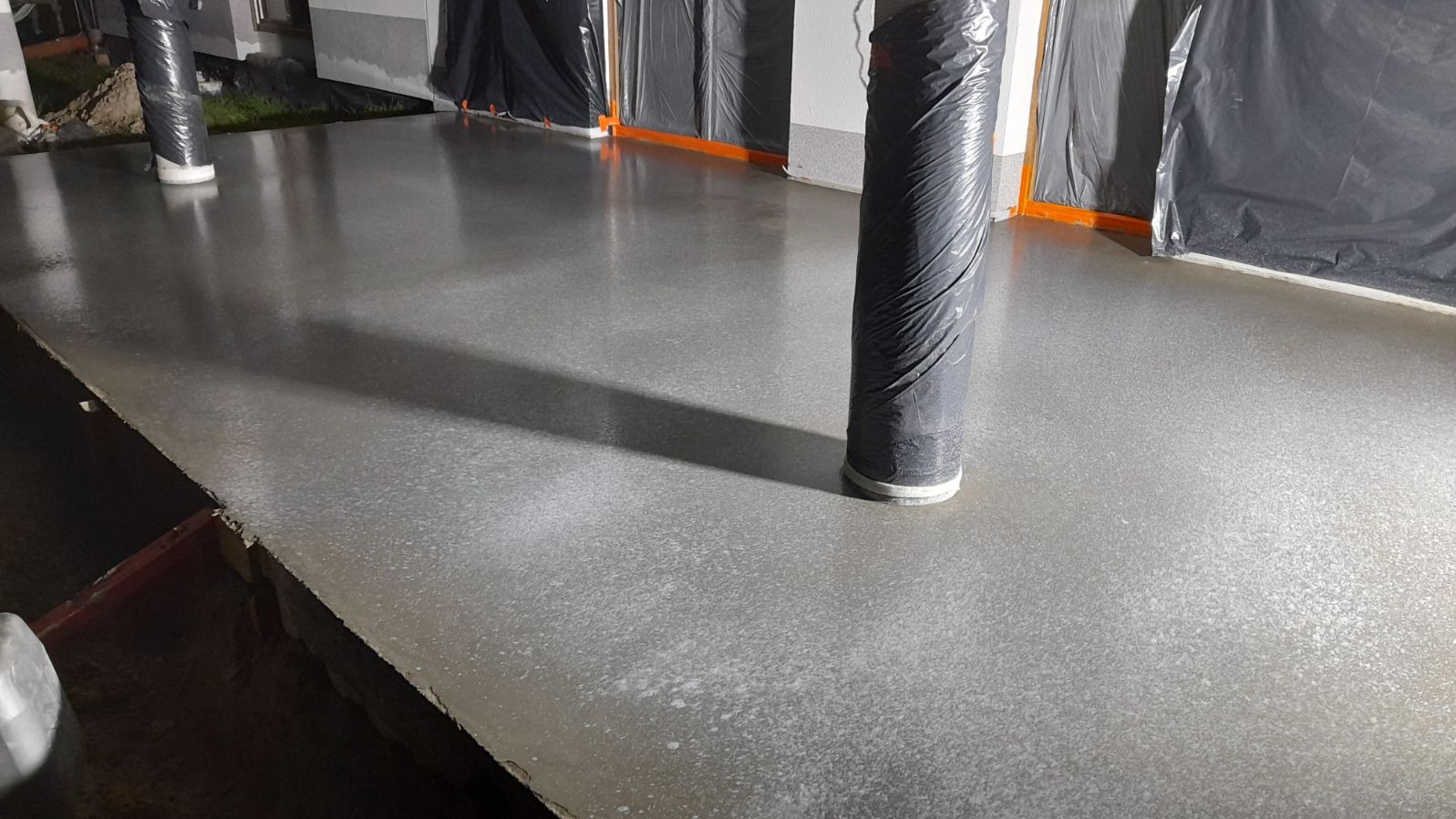 Świeżo wylana, gładka, szara posadzka w pomieszczeniu, z kolumnami zabezpieczonymi folią, częściowo widoczne zabezpieczenia ścian folią malarską.
