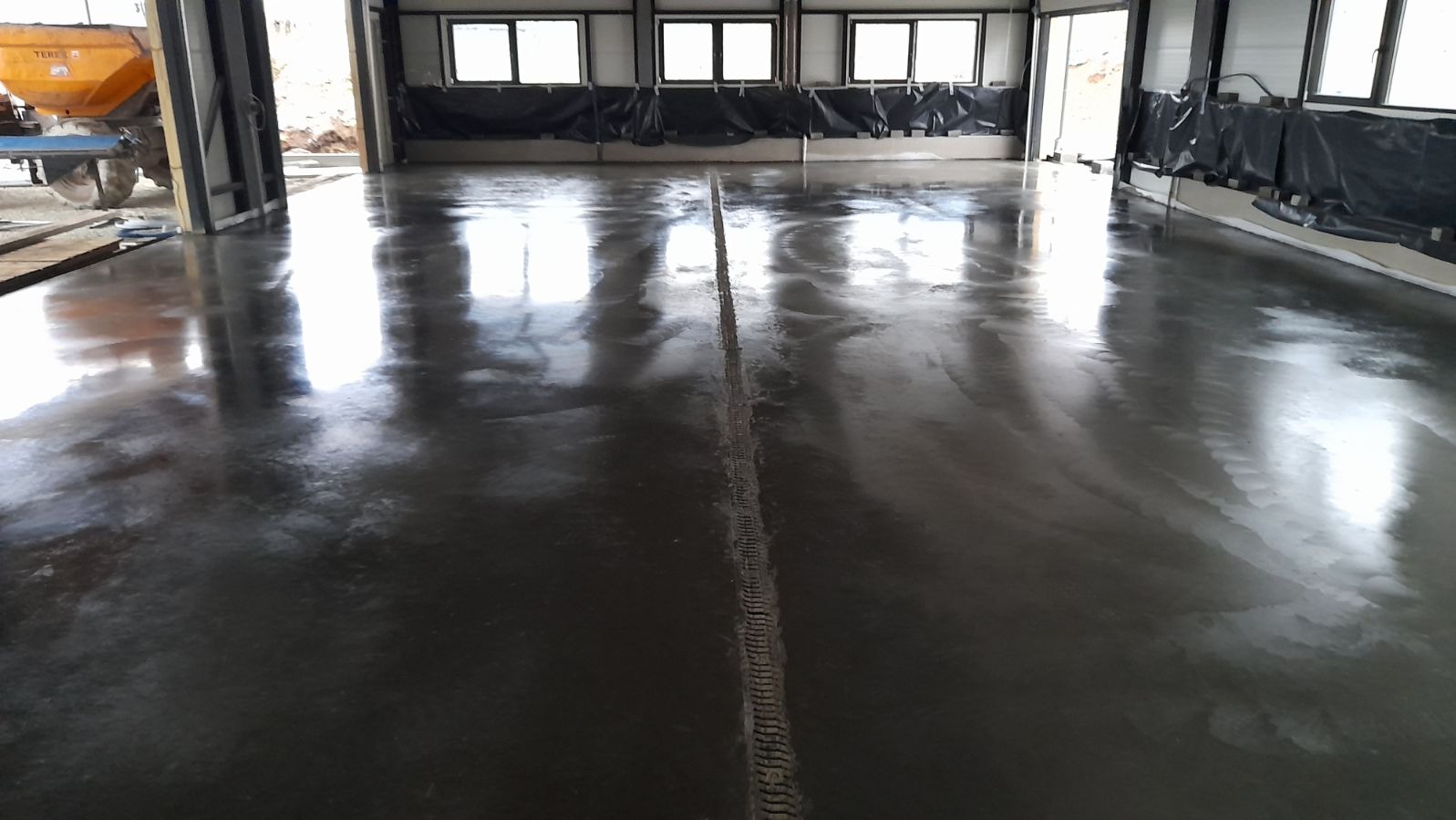 Świeżo wylana, błyszcząca posadzka w hali przemysłowej z widocznymi śladami po oponach maszyny w centralnej części, okna w tle odbijają się w mokrej powierzchni.