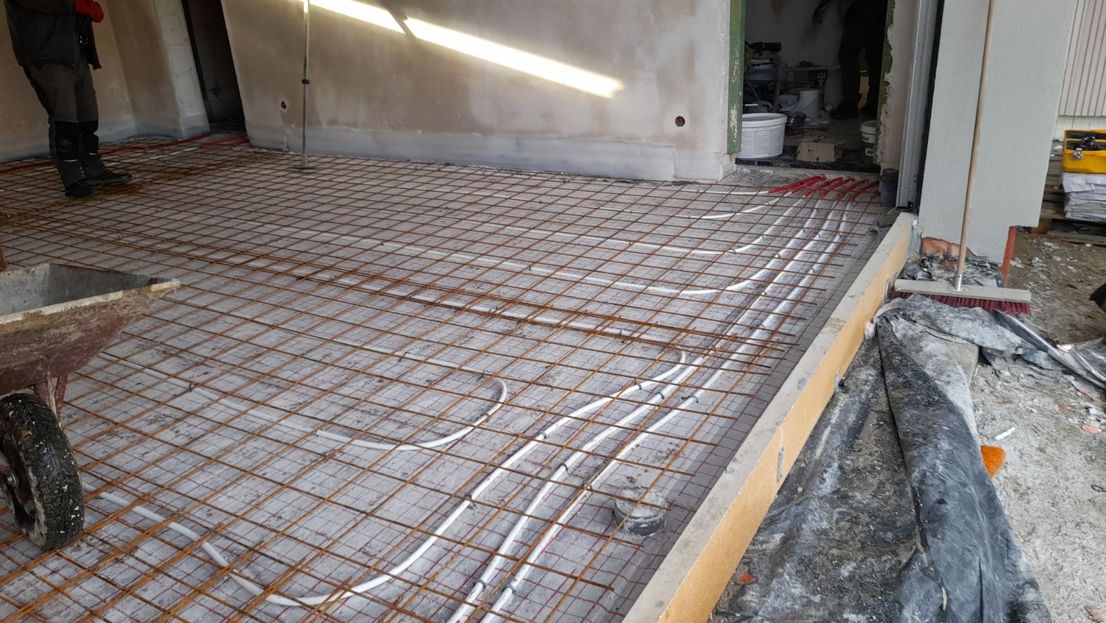 Montaż ogrzewania podłogowego w pomieszczeniu z siatką zbrojeniową, widoczny wózek budowlany i materiały budowlane.