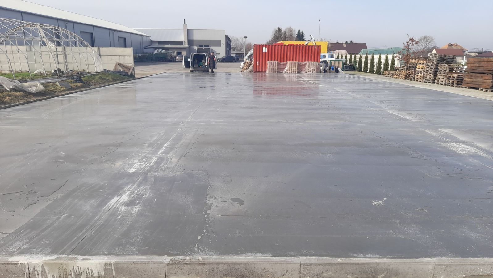 Świeżo wylana, gładka, szara powierzchnia betonowa na zewnątrz, z widocznym vanem i czerwonym kontenerem w tle, sugerującym teren przemysłowy.