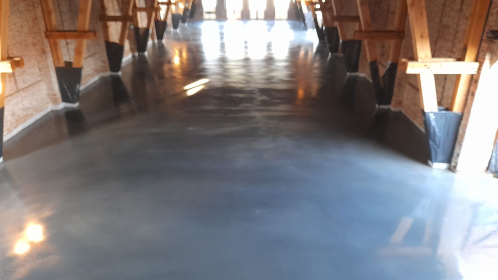 Wypoziomowana, szara posadzka w surowym wnętrzu z drewnianą konstrukcją dachu, zabezpieczoną folią przy ścianach.