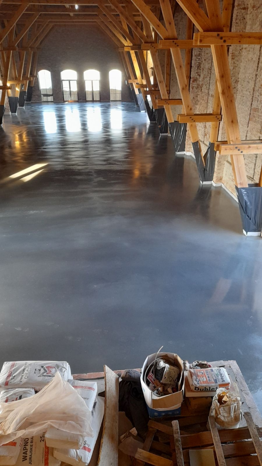 Błyszcząca, szara posadzka w przestronnym, wysokim pomieszczeniu z drewnianą konstrukcją stropu, worki z zaprawą i narzędzia w dolnej części kadru.