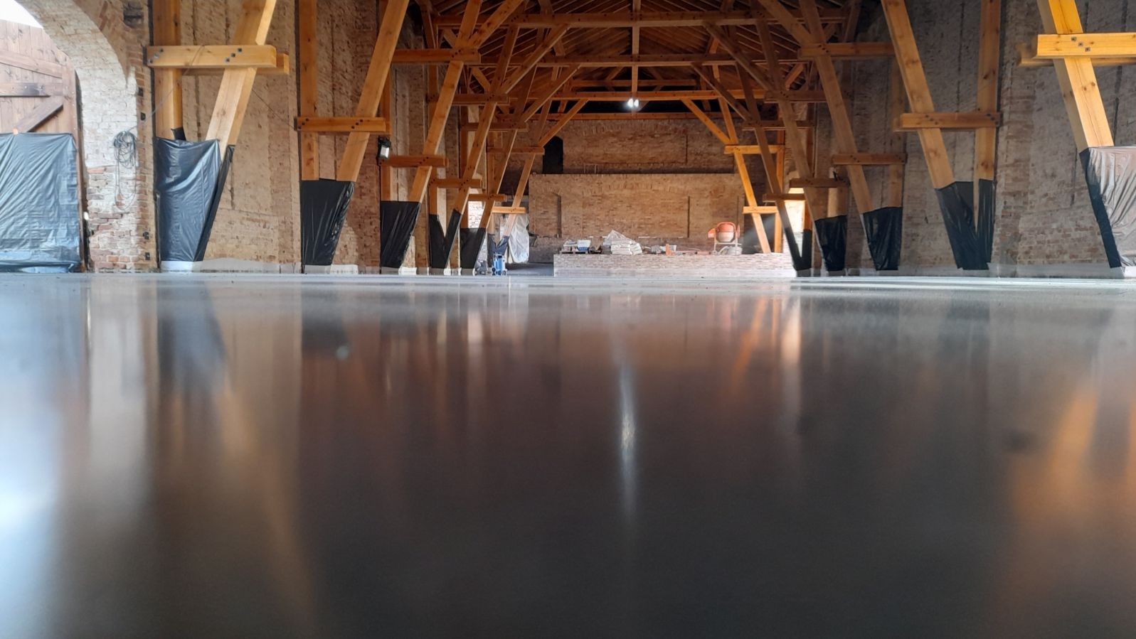 Wypolerowana, szara posadzka przemysłowa w przestronnym, ceglanym wnętrzu z drewnianą konstrukcją stropu, zabezpieczone folią filary, widoczny mikser do betonu w tle.