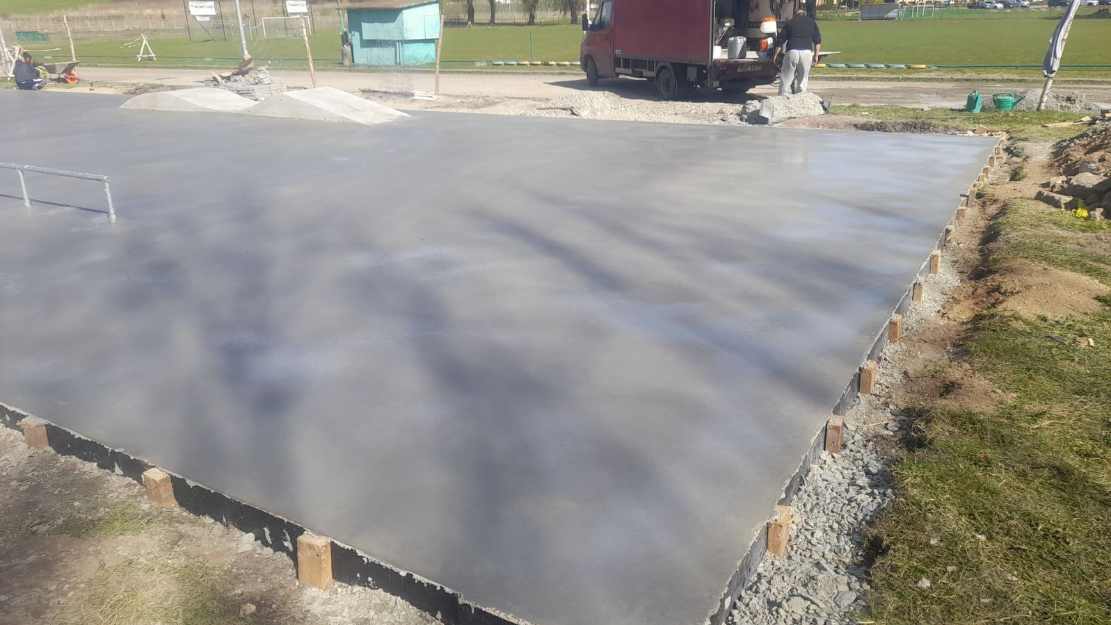 Świeżo wylana, gładka betonowa powierzchnia placu budowy z drewnianym szalunkiem, w tle zaparkowany samochód dostawczy i elementy skateparku.