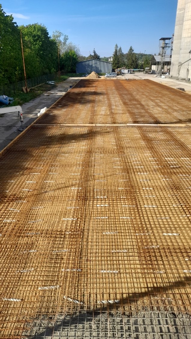 Siatka zbrojeniowa ułożona na piasku, przygotowana pod wylewkę posadzki przemysłowej, widok z góry na dużą powierzchnię roboczą, w tle budynki przemysłowe i drzewa.