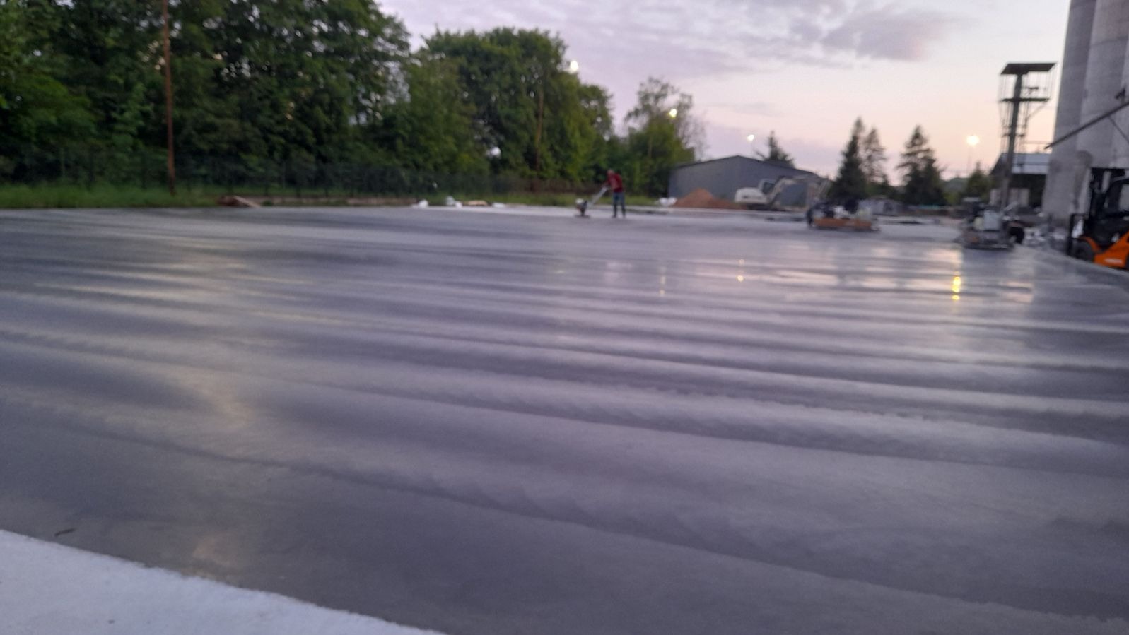 Zacieranie świeżo wylanej betonowej powierzchni przemysłowej na zewnątrz, widoczne maszyny zacierające i pracownik w oddali, w tle budynki przemysłowe i silosy w pochmurny dzień.