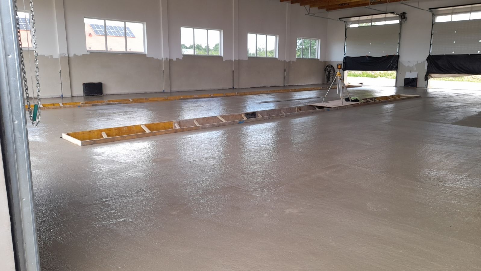 Świeżo wylana, mokra posadzka w hali przemysłowej, z widocznymi drewnianymi szalunkami i laserowym niwelatorem ustawionym na środku, okna w tle.