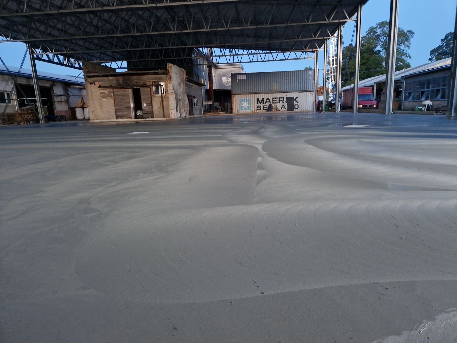 Świeżo wylana, gładka, szara posadzka w hali przemysłowej, z widocznymi śladami po procesie wygładzania, kontenery transportowe w tle.