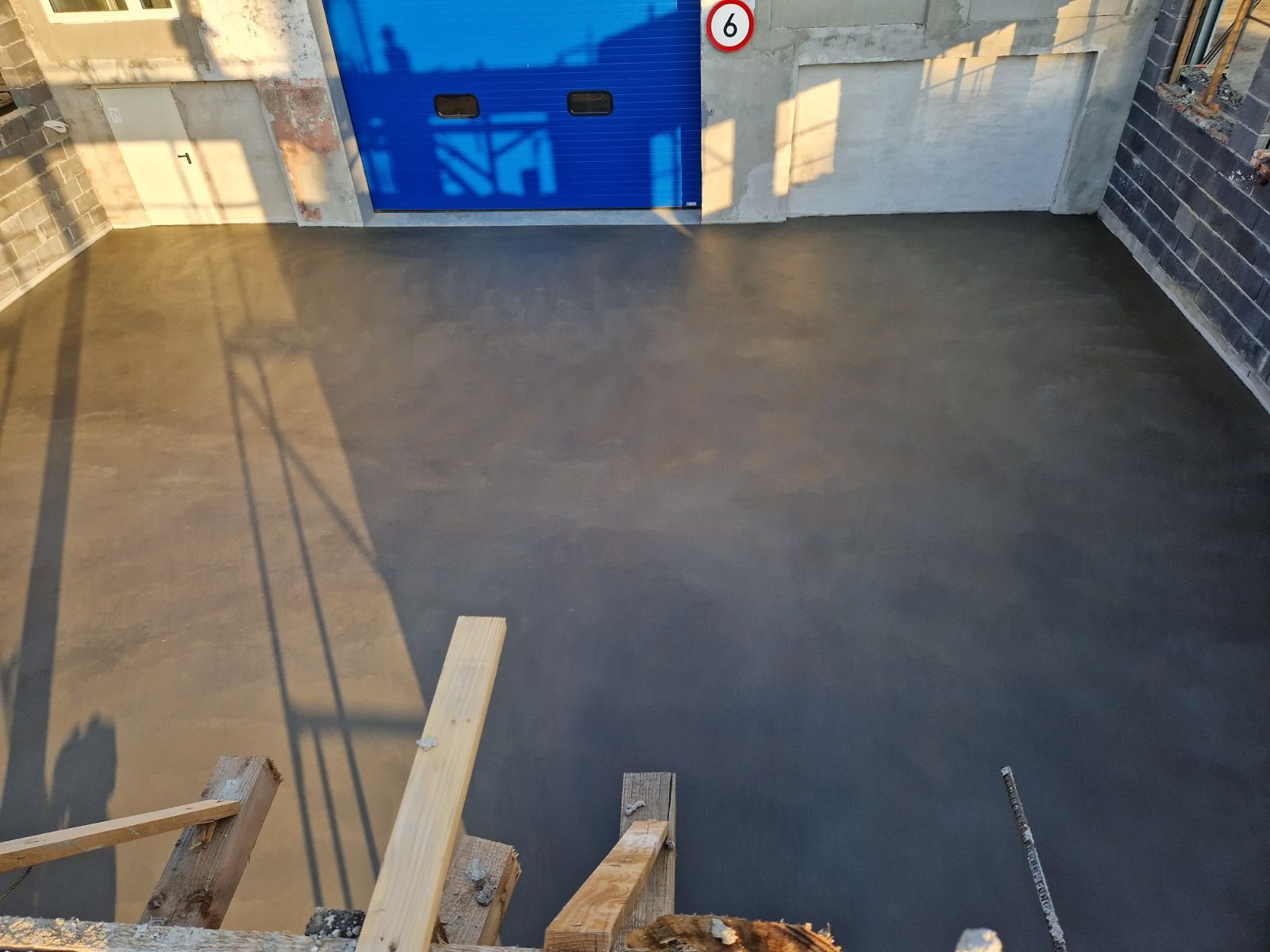 Świeżo wylana, gładka posadzka przemysłowa w hali z niebieską bramą garażową, widziana z góry z fragmentem drewnianego rusztowania w dolnej części kadru.