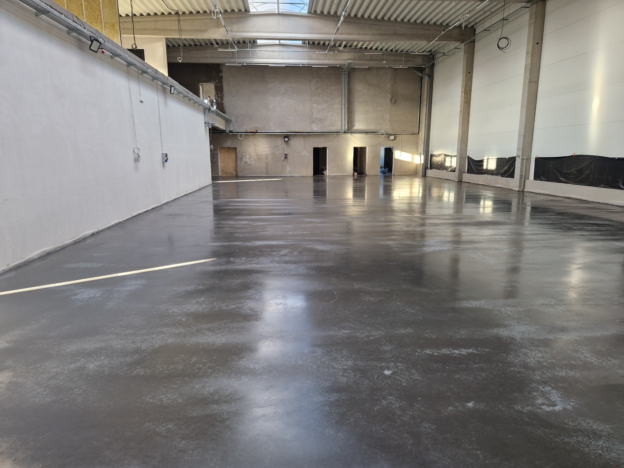 Błyszcząca, szara posadzka przemysłowa w przestronnym, surowym wnętrzu hali z betonowymi ścianami i metalową konstrukcją dachu. Widoczne instalacje pod sufitem i wnęki drzwiowe.