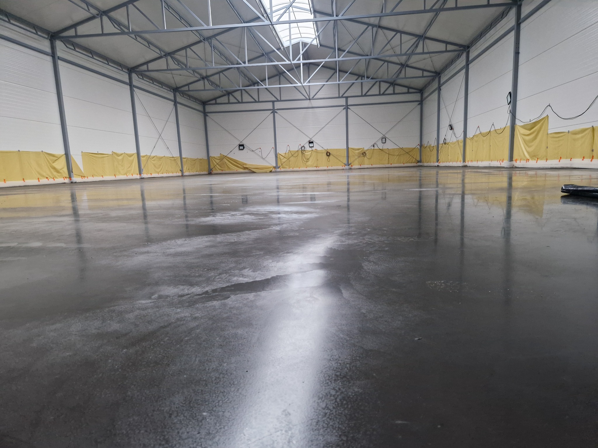Błyszcząca, szara posadzka w hali przemysłowej z żółtymi osłonami na ścianach. Widoczne metalowe elementy konstrukcyjne i świetlik w dachu. Gładka powierzchnia odbija światło.
