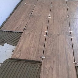 Marek Czarnecki - Proces układania płytek imitujących drewno na podłodze, widoczny klej i krzyżyki dystansowe.