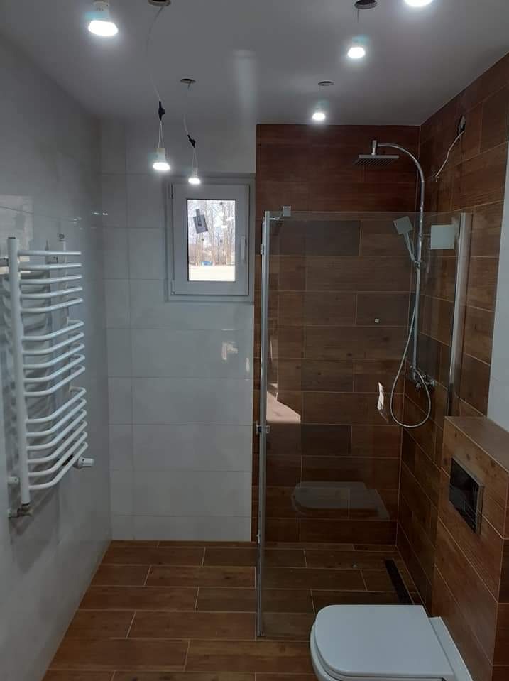 Nowoczesna łazienka z kabiną prysznicową typu walk-in, grzejnikiem drabinkowym, drewnopodobnymi płytkami na ścianach i podłodze oraz oświetleniem punktowym i wiszącym.