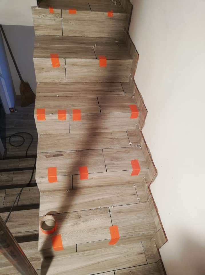 Schody wyłożone płytkami imitującymi drewno, zabezpieczone pomarańczową taśmą malarską, widoczne krzyżyki dystansowe i rolka taśmy.