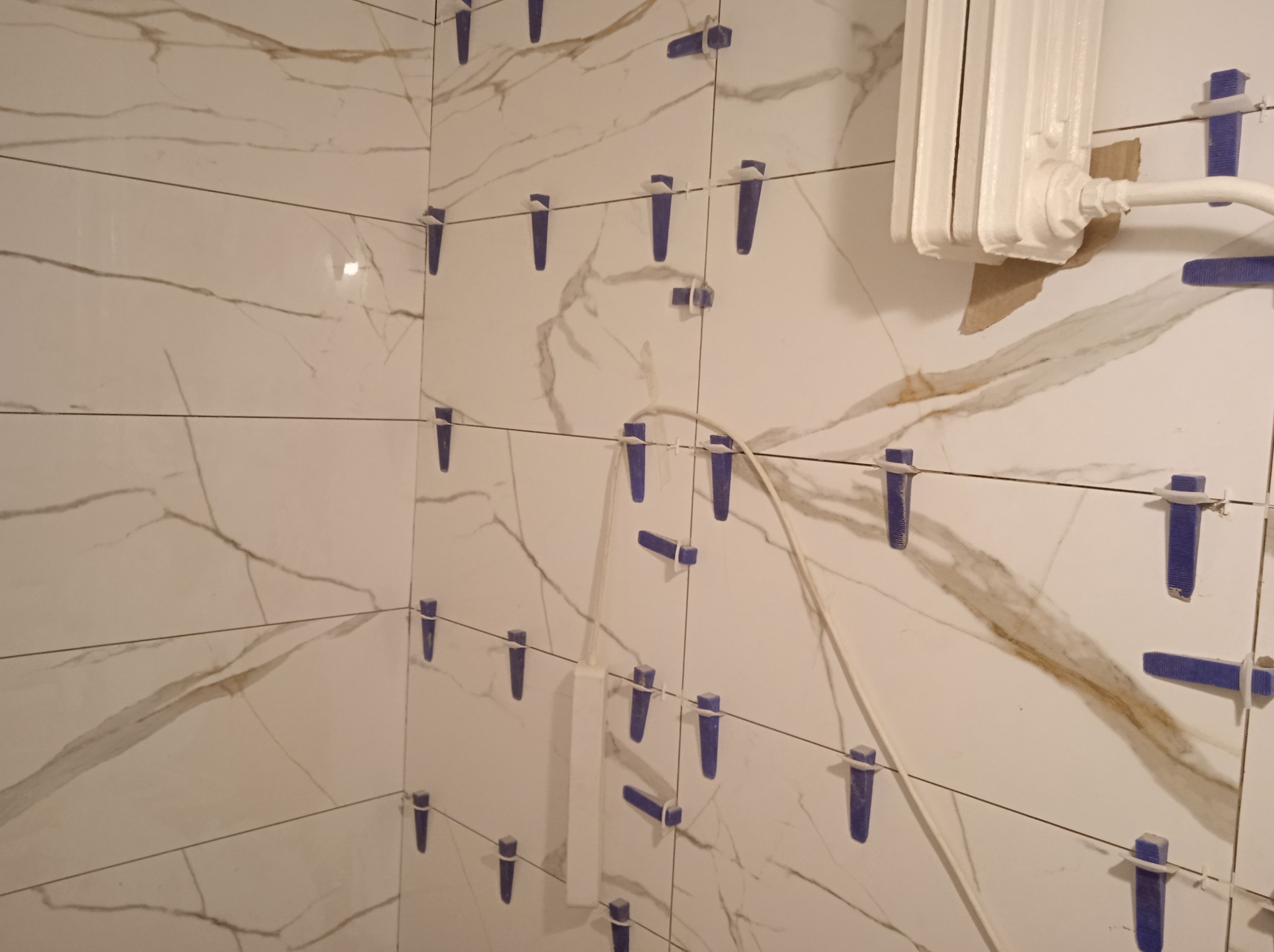 Białe płytki imitujące marmur z widocznymi niebieskimi klinami poziomującymi, grzejnik w tle z widocznymi uszkodzeniami ściany.