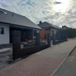 Wznoszenie ogrodzenia z betonowych bloczk&oacute;w w nowoczesnym stylu, widoczne fundamenty i częściowo ułożona kostka brukowa obok posesji.