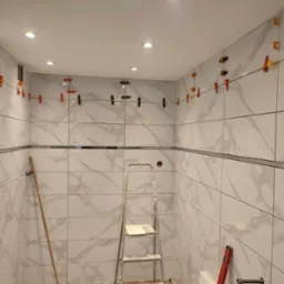 Ściany łazienki wyłożone białymi płytkami imitującymi marmur, z użyciem klipsów poziomujących, drabina w centrum kadru.