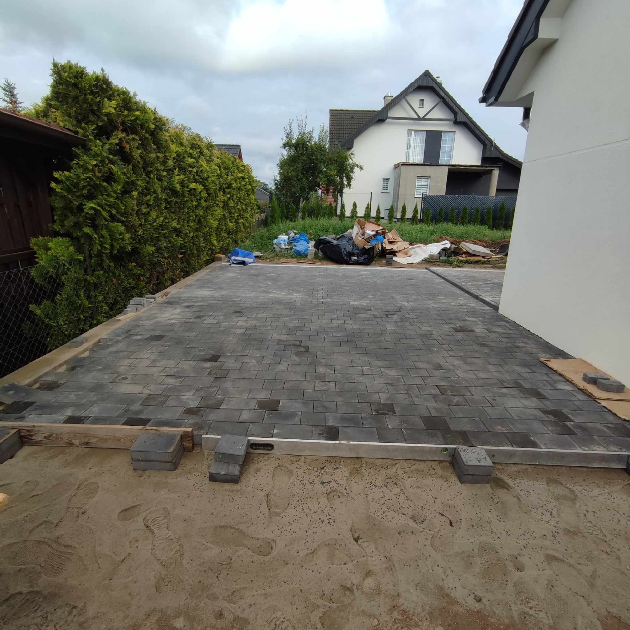 Ułożona częściowo nawierzchnia z szarej kostki brukowej, poziomowana łatą, z widocznymi śladami stóp w podsypce piaskowej i materiałami budowlanymi w tle.